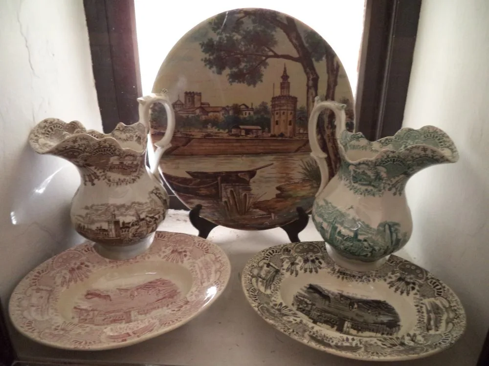 Ceramics exhibited in the naval museum "Torre del Oro" in Seville. In the foreground, there are two Italian-style pitchers and two shaving bowls, all from La Cartuja de Sevilla. In the background, there is a ceramic plate decorated with the Torre del Oro and the Guadalquivir.