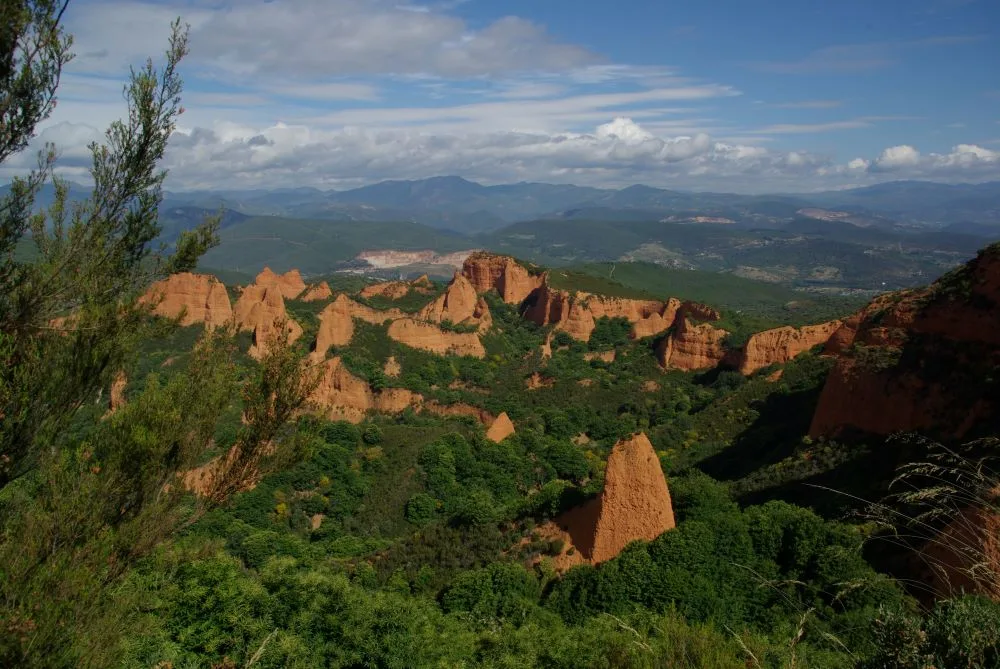 An aerial view of Las Médulas, the ancient Roman gold mine in León, Spain. The landscape features striking red and orange rock formations, with lush green vegetation surrounding the eroded cliffs and peaks. The contrast between the vibrant colors of the terrain and the greenery highlights the historical significance and natural beauty of this UNESCO World Heritage Site.