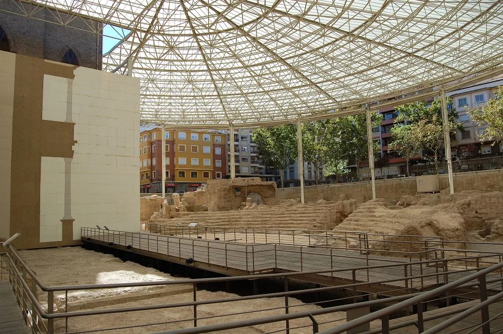 An edited photograph of the Caesaraugusta Roman Theatre Museum in Zaragoza, Spain, showcasing the well-preserved remains of the ancient Roman theatre. The image highlights the semi-circular seating arrangement, stone ruins, and the museum's modern structure integrated with the archaeological site, providing a glimpse into the architectural grandeur of Roman times.