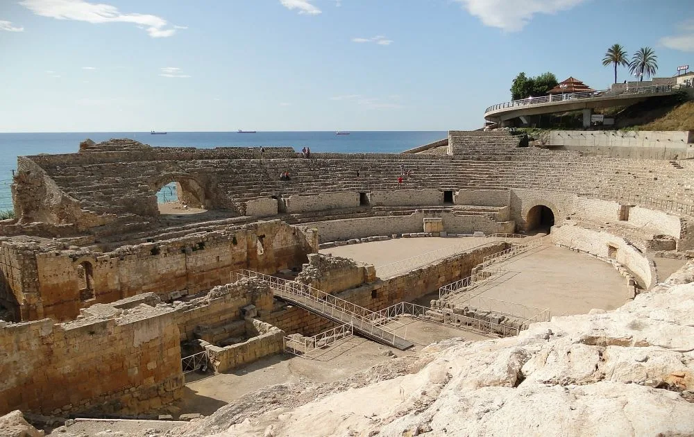 A view of the Roman Amphitheatre of Tarragona in Catalonia, Spain. The ancient stone structure is situated near the Mediterranean coast, with its oval shape and tiered seating partially visible. The photograph highlights the historic architecture against a backdrop of blue sky and scattered clouds, emphasizing the amphitheatre's grandeur and seaside location.