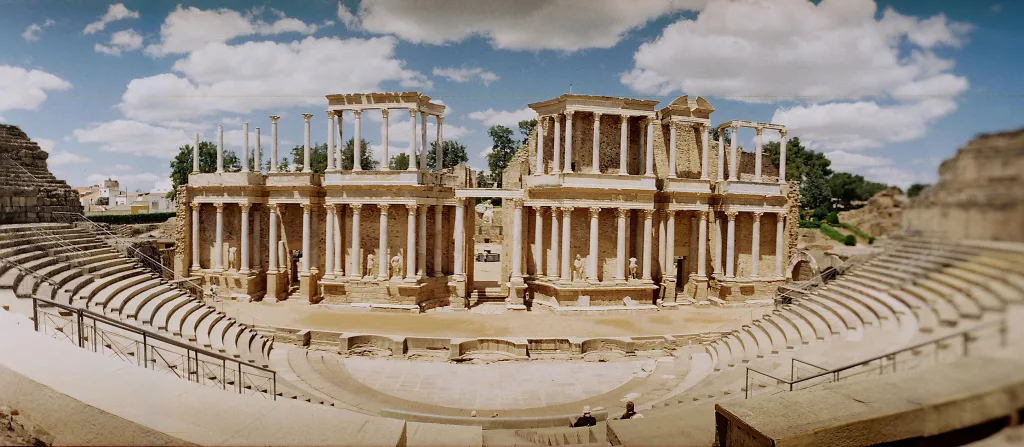 A wide-angle view of the Roman Theatre of Mérida, Spain, showcasing the well-preserved ancient structure with its grand semicircular seating and elaborately decorated stage backdrop. The photograph highlights the intricate stonework and architectural design against a backdrop of a clear sky. Edited for enhanced clarity and visual impact.