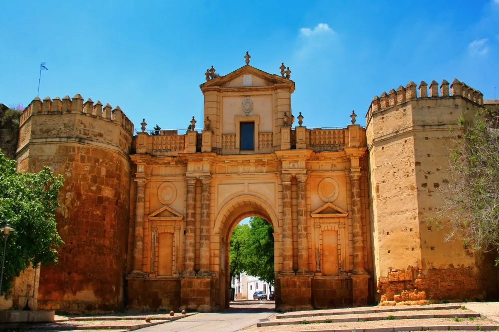 A scenic view of Carmona, Seville, Spain, showcasing historical architecture with warm-toned buildings, a prominent tower, and lush greenery in the background under a clear sky.