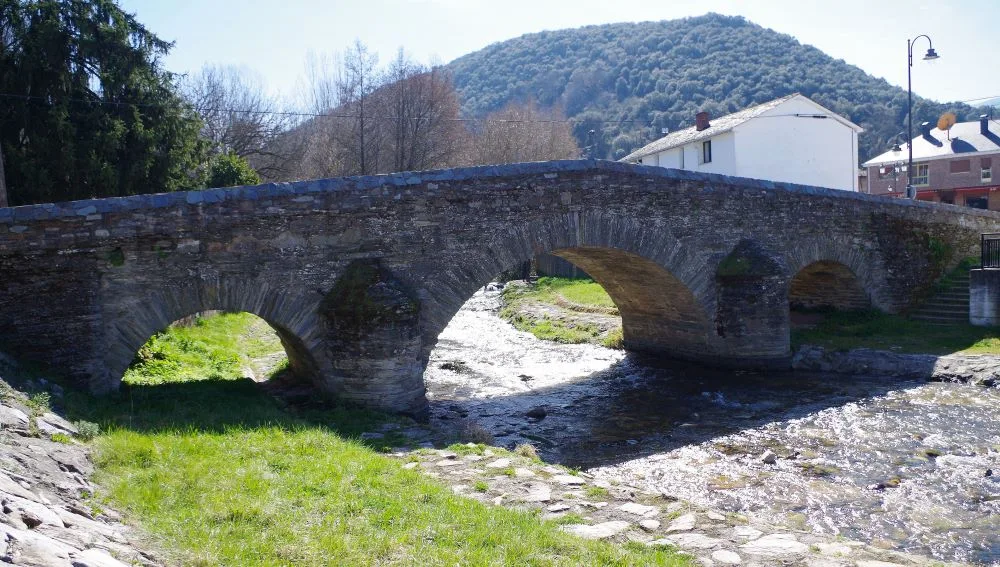 A medieval stone bridge in Toral de Merayo, Ponferrada, Spain, gracefully arching over the Oza River. Surrounded by lush greenery, the historic structure blends harmoniously with the natural landscape, evoking a sense of timeless tranquility in the Bierzo region.