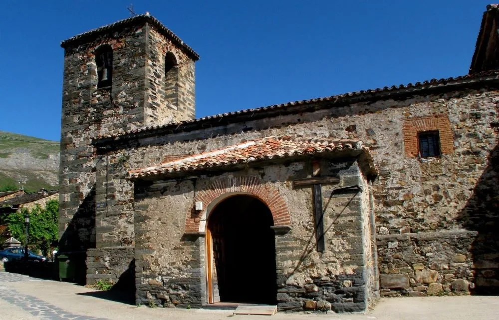San Ildefonso Church in Valverde de los Arroyos, Spain, showcasing its traditional black slate architecture with a bell tower rising against a clear blue sky.