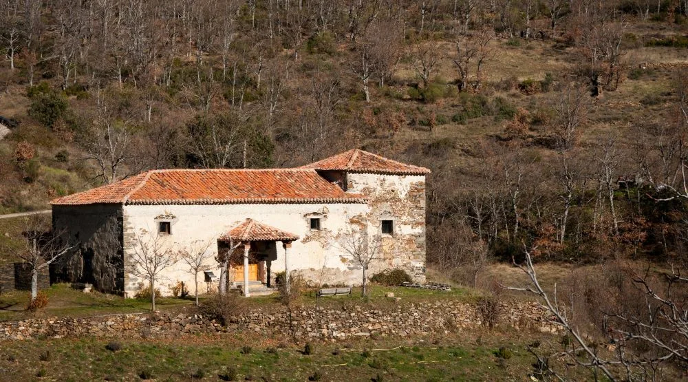 A small stone hermitage, the Hermitage of the Virgen de Gracia, nestled in the serene landscape of Valverde de los Arroyos, Spain. The rustic architecture blends with the surrounding nature, creating a peaceful and timeless scene.