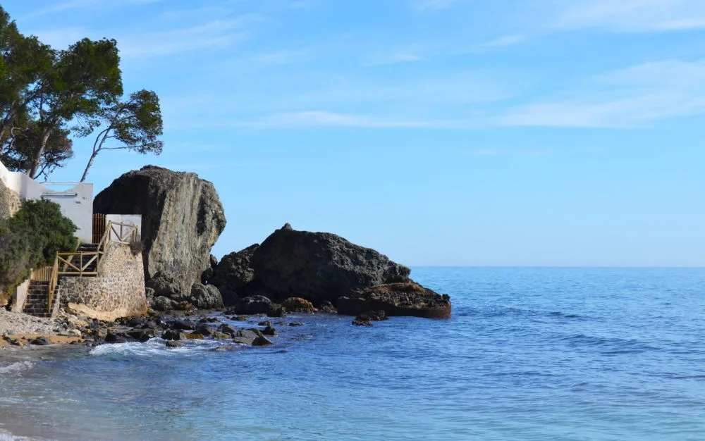A coastal view of Cap Negret in Altea, Spain, featuring a rocky shoreline, blue Mediterranean waters, and a clear sky.