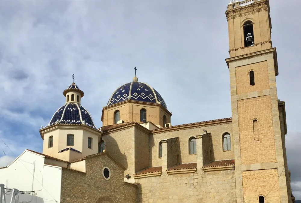 A view of Església de la Mare de Déu del Consol in Altea, Spain, showcasing its iconic blue-tiled domes and Mediterranean architectural style against a clear sky.