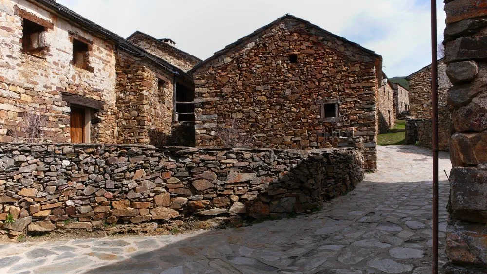 A scenic view of Valverde de los Arroyos, Spain, showcasing the traditional black slate architecture of the village set against a mountainous backdrop. The rustic charm of the stone houses and narrow streets reflects the region’s rich cultural heritage.