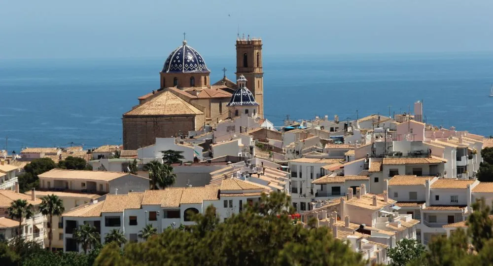 A scenic view of the Església de la Mare de Déu del Consol in Altea, Spain, showcasing its distinctive blue-tiled domes and Mediterranean architectural charm against a bright sky.